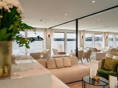 Helle, moderne Schiffslounge auf der AMADEUS Nova mit Panoramafenstern und Blick auf den Fluss. Gemütliche Sitzgruppen in hellen Farben.