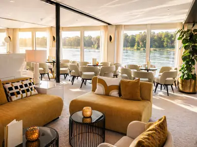 Helle und geräumige Lounge auf der AMADEUS Nova. Gemütliche Sofas, Sessel, kleine Tische. Große Fenster bieten weiten Blick auf den sonnigen Fluss und bewaldete Ufer.