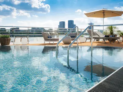 Ein luxuriöser Pool auf der AMADEUS Nova mit Edelstahltreppe auf einem sonnigen Schiffsdeck, umgeben von eleganten Loungemöbeln und Pflanzen, mit einer Stadt-Skyline am Horizont.