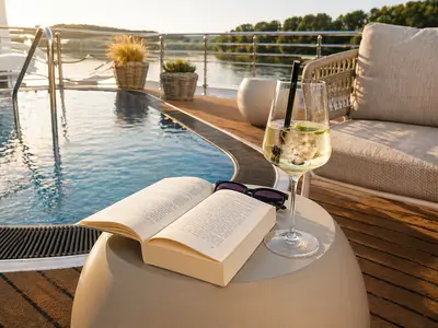 Offenes Buch und Cocktail auf Beistelltisch am Poolrand auf dem Schiffsdeck der AMADEUS Nova. Sonnenuntergangslicht, Lounge-Sessel und Ufervegetation im Hintergrund.