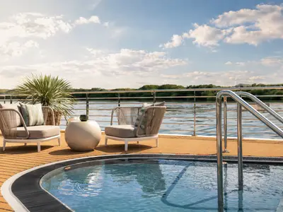 Whirlpool auf einem Holzdeck der AMADEUS Nova. Moderne Lounge-Sessel, Beistelltisch und Pflanze umgeben ihn. Im Hintergrund ein Fluss mit bewaldeten Ufern unter blauem Himmel mit Wolken.