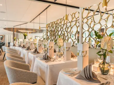 Ein elegantes Restaurant auf der AMADEUS Nova mit langen Reihen festlich gedeckter Tische. Weiße Tischdecken, Servietten, Gläser und Blumendekorationen dominieren den Raum. Eine goldene, durchbrochene Raumteiler-Wand trennt die Bereiche.