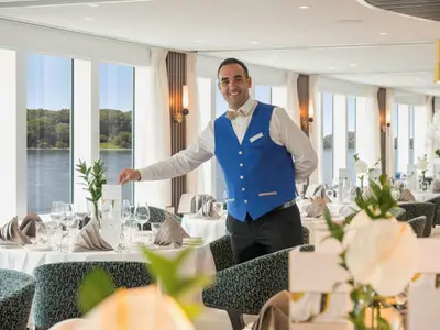 Ein lächelnder Kellner in blauem Gilet steht in einem eleganten Schiffsrestaurant. 