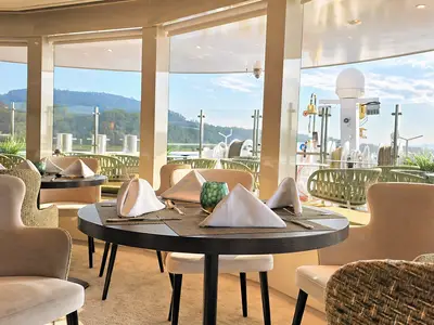 Innenansicht eines Restaurants mit elegant gedeckten Tischen und bequemen Stühlen. Große Panoramafenster bieten einen weiten Blick auf grüne Hügel und das Wasser. 