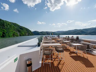 Blick über das Sonnendeck der AMADEUS Nova mit Gästen an Tischen, umgeben von einem Fluss und grünen Bergen unter blauem Himmel.