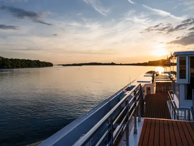 Von der AMADEUS Nova aus blickt man auf einen ruhigen Fluss. Die Sonne geht hinter bewaldeten Ufern unter und taucht die Szene in warmes Licht.