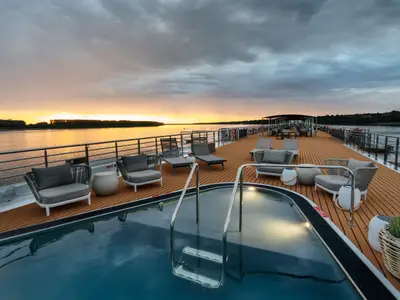 Luxuriöses Sonnendeck der AMADEUS Nova bei Sonnenuntergang. Ein ovaler Pool, moderne Lounge-Sessel und Tische sind zu sehen. Die Sonne spiegelt sich im ruhigen Flusswasser am Horizont.
