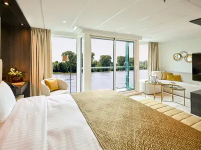 Luxuriöse Flusskreuzfahrt-Suite auf der AMADEUS Nova mit Doppelbett, elegantem Wohnbereich und Panoramafenstern. Blick auf den Fluss und grüne Ufer. Ein Sessel, TV, Champagner und Gebäck sind sichtbar.