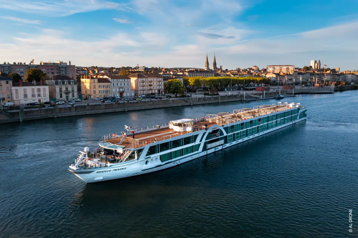 Blick aus der Vogelperspektive auf das Flusskreuzfahrtschiff AMADEUS Provence auf einem breiten Fluss. Im Hintergrund eine historische Stadtkulisse mit Gebäuden und Kirchtürmen unter blauem Himmel.