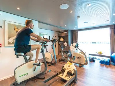 Ein Mann in sportlicher Kleidung trainiert auf der AMADEUS Provence auf einem Heimtrainer in einem modernen Fitnessraum an Bord eines Kreuzfahrtschiffs. Im Hintergrund sind weitere Fitnessgeräte, gerollte Handtücher und ein Fenster mit Blick nach draußen zu sehen.