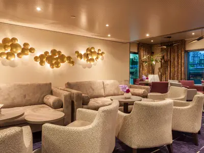 Eine moderne und elegante Lounge auf der AMADEUS Provence mit beigen Sofas, gemusterten Sesseln und runden, goldenen Wandleuchten, die ein warmes Licht abgeben. Im Hintergrund sind Fenster mit Blick nach draussen zu sehen.