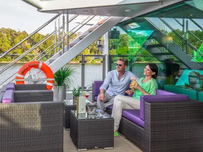 Ein Mann und eine Frau sitzen auf dem Loungedeck der AMADEUS Provence, trinken Cocktails und blicken auf den Fluss und die bewaldeten Ufer. 