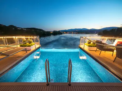 Ein Infinity-Pool auf dem Außendeck der AMADEUS Provence bei Abenddämmerung. Das türkise Wasser des Pools ist beleuchtet und spiegelt den klaren Abendhimmel wider. Links und rechts des Pools befinden sich Bereiche mit Holzdecks, Liegestühlen und Pflanzen in Kübeln, umrahmt von Lichterketten. 