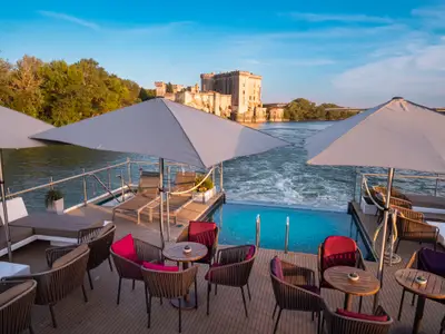 Sonnendeck der AMADEUS Provence mit Infinity-Pool, Lounge und Schirmen. Blick auf Fluss, historisches Schloss und bewaldete Ufer.