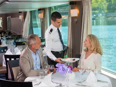 Ein Kellner in Uniform serviert einem Laecheln einem Paar am Tisch ein Dessert, waehrend sie auf der AMADEUS Provence speisen und dabei auf das vorbeiziehende Wasser blicken.