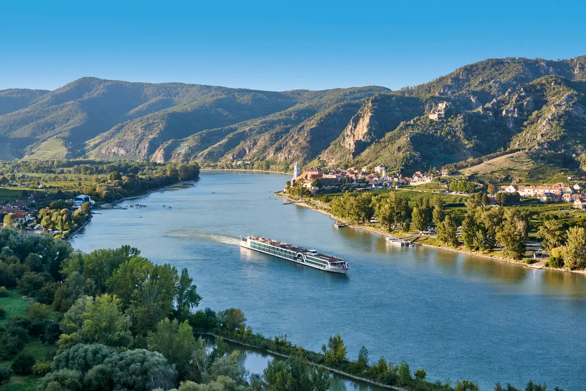 Eine Vogelperspektive zeigt die AMADEUS Queen auf einem breiten Fluss, der sich durch eine grüne Berglandschaft schlängelt. Am Ufer sind kleine Dörfer und Weinberge zu sehen.