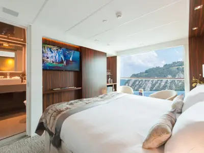 Eine luxuriöse Kreuzfahrtkabine auf der AMADEUS Queen mit Doppelbett, hellem Badezimmer, Flachbildfernseher und raumhoher Fensterfront mit Blick auf bewaldete Berge und ruhiges Wasser.