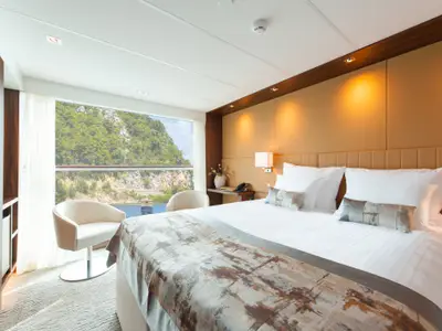 Helle, luxuriöse Außenkabine auf der AMADEUS Queen. Ein grosses Doppelbett, zwei Sessel und ein Tisch sind zu sehen. Die Panoramafenster bieten Blick auf grüne Berge und Wasser.