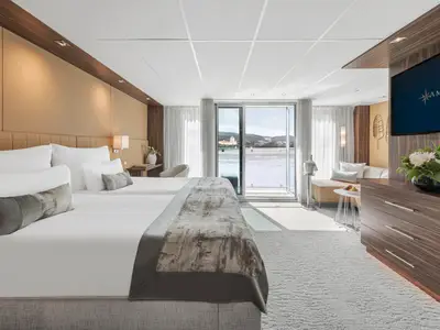 Geräumige Suite auf der AMADEUS Queen mit zwei Einzelbetten, Sofa, TV und Balkon mit Panoramablick auf Fluss und Uferlandschaft bei Tageslicht.