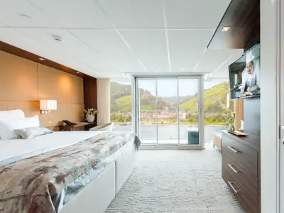 Eine helle, moderne Suite an Bord der AMADEUS Queen mit einem gemachten Bett, einem Fernseher und einer großen Glasfront mit Blick auf einen Fluss und grüne Berge mit einer Burg.