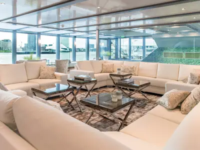 Helle, moderne Lounge an Bord der AMADEUS Queen mit cremefarbenen Sofas, mehreren Couchtischen und grossen Fenstern, die den Blick auf eine städtische Uferpromenade freigeben.