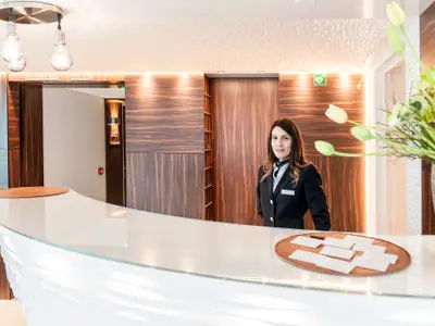 Eine weibliche Crew-Mitarbeiterin in Uniform lächelt auf der AMADEUS Queen hinter einer modernen Rezeption aus Glas und Holz. 