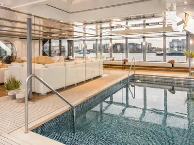 Luxuriöser Innenpool mit Mosaikfliesen und angrenzendem Loungebereich auf der AMADEUS Queen. Grosse Fenster bieten Blick auf eine Stadt am Wasser. 
