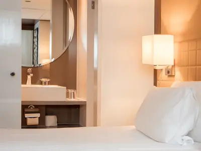 Eine modern eingerichtete Kreuzfahrtkabine auf der AMADEUS Queen zeigt eine weisse Nachttischlampe, ein gepolstertes Kopfteil, und einen Spiegelschrank über einem Waschbecken. Weisse Kissen liegen auf dem Bett.