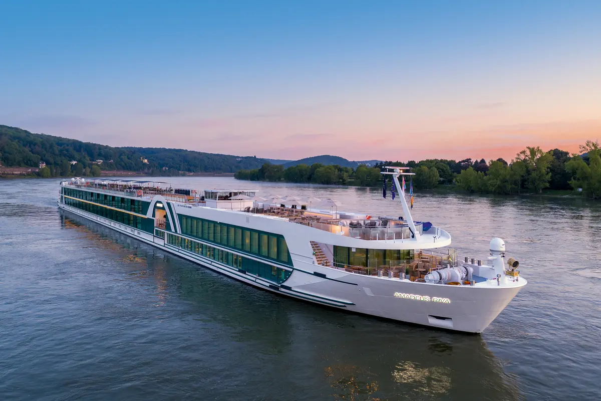 Elegantes Flusskreuzfahrtschiff AMADEUS Riva bei Sonnenuntergang auf einem Fluss, umgeben von grünen Ufern. Die warmen Lichter des Schiffes spiegeln sich im Wasser.