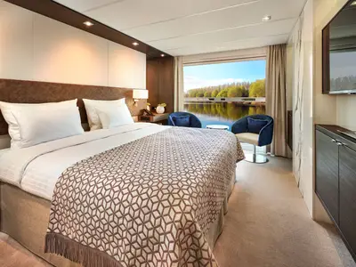 Eine helle und moderne Kabine auf der AMADEUS Riva. Ein grosses Doppelbett mit weissen Laken und einer braun gemusterten Tagesdecke befindet sich links. Rechts davon ist ein Panoramafenster mit Blick auf einen ruhigen Fluss und bewaldete Ufer zu sehen. Zwei blaue Sessel und ein kleiner Tisch stehen am Fenster. 