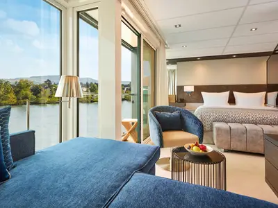 Innenansicht einer luxuriösen Suite auf der AMADEUS Riva. Ein bequemes Sofa, Sessel und ein Doppelbett sind zu sehen. Der Blick durch die große Glasfront fällt auf einen ruhigen Fluss und bewaldete Ufer mit Hügeln unter blauem Himmel.
