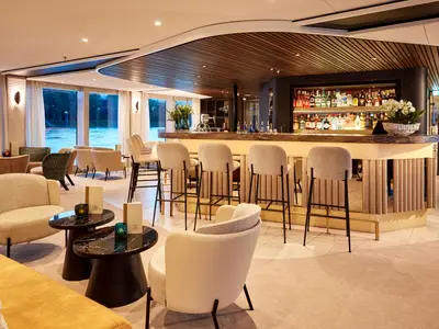 Eine stilvolle Bar-Lounge auf der AMADEUS Riva mit einer langen Bar, Barhockern, Sesseln und großen Panoramafenstern mit Blick auf einen Fluss und die Ufervegetation.