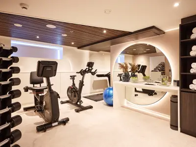 Ein gut ausgestatteter Fitnessraum an Bord der AMADEUS Riva. Links ein Hantelständer, in der Mitte zwei Fitnessfahrräder und ein Laufband. Rechts ein großer runder, beleuchteter Spiegel über einer Theke mit frischem Obst und Getränken. 