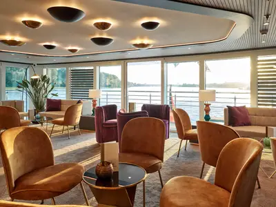 Eine moderne und gemuetliche Lounge an Bord der AMADEUS Riva. Mehrere braune und lila Sessel sowie Sofas sind auf einem Teppichboden arrangiert. 