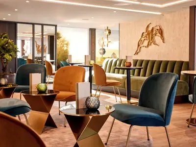 Eine moderne und elegante Lounge auf der AMADEUS Riva. Mehrere goldene und dunkelblaue Samtsessel umgeben kleine Tische. Im Hintergrund eine olivgruene geraffte Sitzbank. 