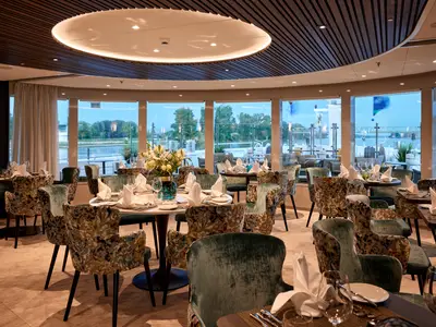 Ein elegantes Restaurant auf der AMADEUS Riva mit vielen Tischen, die für ein Abendessen gedeckt sind, und grossen Fenstern, die den Blick auf einen Fluss und Bäume freigeben.
