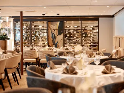 Ein elegantes Restaurant auf der AMADEUS Riva mit einer grossen Glasvitrine voller Weinflaschen im Hintergrund. Mehrere Tische sind weiss gedeckt und mit grauen Stoffservietten und Weinglaesern versehen. Bequeme beige und graue Sessel stehen um die Tische.
