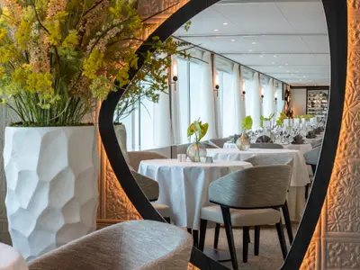 Ein elegantes Restaurant auf der AMADEUS Riva, gesehen durch einen Spiegel. Weisse Tische sind gedeckt, Pflanzen schmuecken den Raum, grosse Fenster bieten Ausblick. 