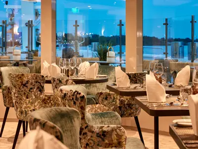 Elegantes Restaurant auf der AMADEUS Riva bei Abendstimmung. Die Tische sind gedeckt mit Besteck, Gläsern und weißen Servietten. Gemusterte und samtbezogene Stühle dominieren den Vordergrund. Große Fenster bieten einen Blick auf das ruhige Wasser und eine beleuchtete Uferlandschaft am Horizont.