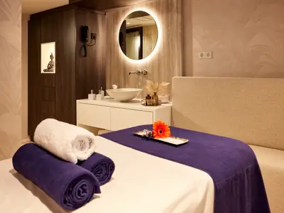 Heller Spa-Behandlungsraum auf der AMADEUS Riva mit violett-weisser Massageliege im Vordergrund. Dahinter ein Waschbeckenbereich mit beleuchtetem Spiegel und eine Buddha-Statue in einer beleuchteten Nische.