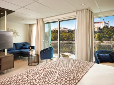 Moderne Flusskreuzfahrt-Suite auf der AMADEUS Riva mit Doppelbett, blauer Sitzecke, TV und Balkon. Blick auf einen Fluss, bewaldete Ufer und eine helle Burg auf einem Hügel.