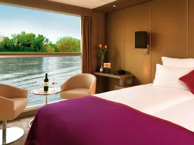 Helle Flusskreuzfahrtkabine auf der AMADEUS Silver II mit Panoramafenster. Blick auf Fluss und bewaldete Ufer. Doppelbett, zwei Sessel, Beistelltisch mit Wein und Gebäck.