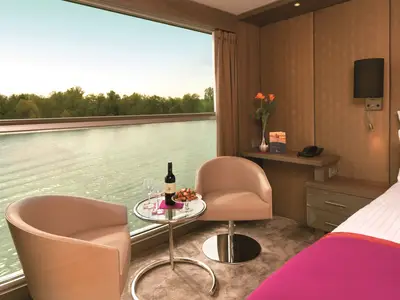 Eine helle, gemütliche Aussenkabine auf der AMADEUS Silver II mit großem Panoramafenster. Zwei Sessel, ein Glastisch mit Rotwein und Snacks stehen am Fenster. Draußen ist ein Fluss mit bewachsenen Ufern sichtbar. Ein Bett und Nachttisch sind rechts zu sehen.