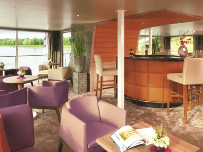 Eine helle Bar-Lounge auf der AMADEUS Silver II. Grosse Fenster bieten Blick auf einen Fluss und grüne Ufer. Lila Sessel und holzfarbene Tische füllen den Raum, eine braune Bar mit weissen Barhockern steht im Hintergrund.