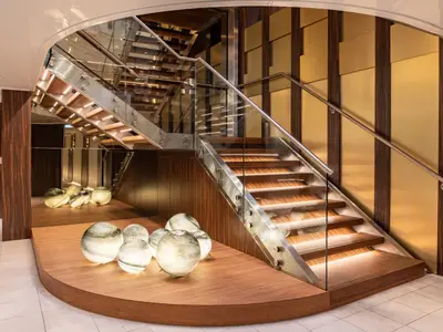 Moderne, offene Treppe in einem Schiffsatrium auf der AMADEUS Star mit beleuchteten Holzstufen, Glasgeländer und Edelstahlrahmen. Dunkle Holzwände mit goldenen Paneelen und leuchtende Glaskugeln auf einem Podest schaffen eine elegante Atmosphäre.