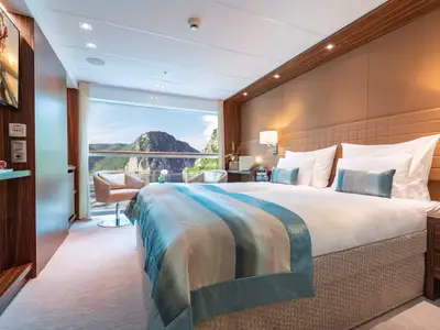 Luxuriöse Kreuzfahrtschiff-Kabine auf der AMADEUS Star mit Doppelbett und blauer Tagesdecke. Große Fensterfront mit Panoramablick auf eine beeindruckende Berg- und Flusslandschaft.
