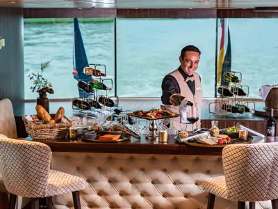 Ein lächelnder Barkeeper auf der AMADEUS Star gießt Rotwein in ein Glas an einer reich gedeckten Bar mit Snacks und Flaschen.
