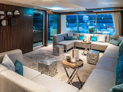 Elegante Kreuzfahrtschiff-Lounge auf der AMADEUS Star mit beigen Sofas, blauen Kissen und modernen Tischen. Große Fenster bieten Flussblick bei Abendstimmung. Eine Holzwand mit runden Spiegeln ist links.