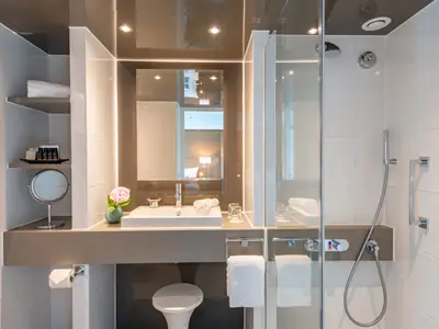 Modernes, helles Badezimmer einer Kreuzfahrtkabine auf der AMADEUS Star mit Waschbecken, Dusche, weissen Fliesen und Kosmetikprodukten. Der Spiegel zeigt den Schlafbereich der Kabine.