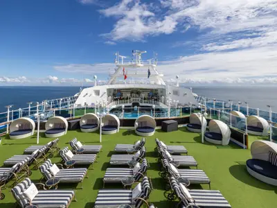 Großes Sonnendeck der Carnival Adventure mit Liegestühlen, runden Tagesbetten auf grünem Kunstrasen und einem Pool. Im Hintergrund sind das Meer und der blaue Himmel zu sehen.